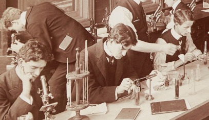 A black-and-white photograph of men in suits seated at a long table, where they are looking through microscopes and titrating substances in test tubes