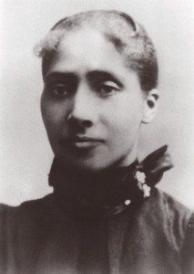 Black and white portrait photograph of Emma Reynolds, straight-faced with a steady gaze. She is dressed in a black dress with a fancy collar, and her hair is up.