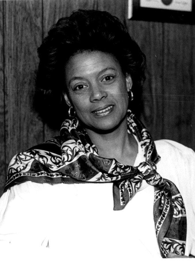 Black woman in white clothes and silk scarf around her neck smiling at the camera