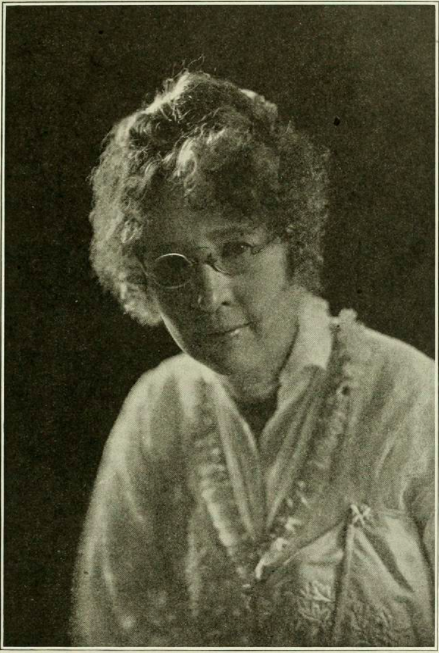 Middle aged white woman wearing glasses and white embellished clothes, posing for her portrait 