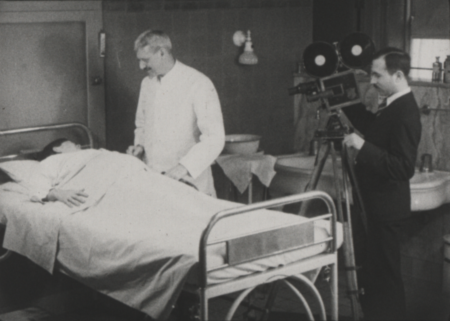 Male Doctor in White coat attending to a woman in labor. Man in a black suit to the right filming the process.